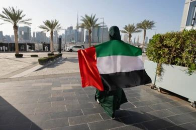الاحتفال بالاعياد الوطنية للشعوب، لاحتفال بذكرى استقلال دولة الإمارات العربية المتحدة،صورة من الخلف لامراة عربية خليجية اماراتية ترتدي العباءة والحجاب تضع علم الإمارات على كتفيها في النهار، التجول في الشوارع بايماءات السعادة والفخر والاعتزاز، مفهوم حب الوطن والانتماء اليه