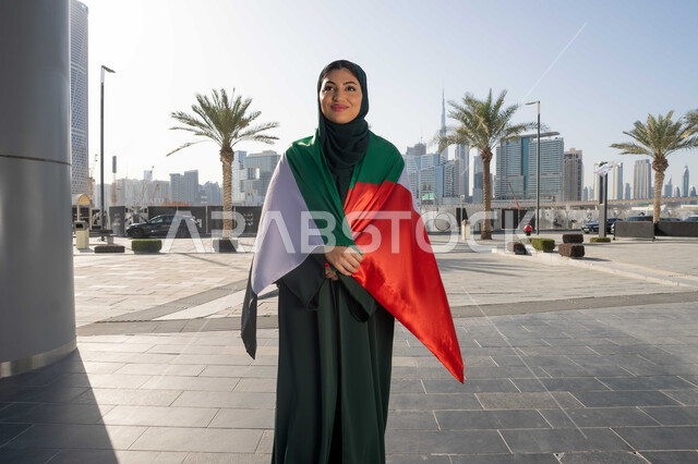 إحياء ذكرى استقلال البلاد، اليوم الوطني الإماراتي 2 ديسمبر، امراة عربية خليجية اماراتية ترتدي العباءة والحجاب تضع علم الإمارات على كتفيها في النهار، التجول في الشوارع بايماءات السعادة والفخر والاعتزاز، مفهوم حب الوطن والانتماء اليه