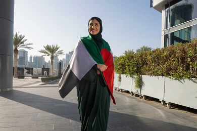 مفهوم حب الوطن والانتماء اليه، اليوم الوطني الإماراتي 2 ديسمبر، الاحتفال بذكرى استقلال دولة الإمارات العربية المتحدة، امراة عربية خليجية اماراتية ترتدي العباءة والحجاب تضع علم الإمارات على كتفيها في النهار، التجول في الشوارع بايماءات السعادة والفخر والاعتزاز