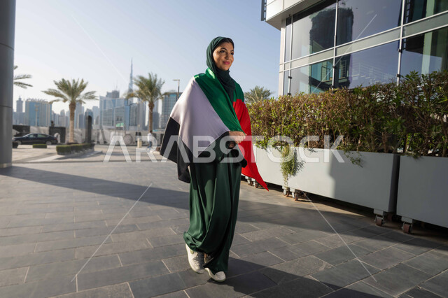 الاحتفال بذكرى استقلال دولة الإمارات العربية المتحدة، اليوم الوطني الإماراتي 2 ديسمبر، امراة عربية خليجية اماراتية ترتدي العباءة والحجاب تضع علم الإمارات على كتفيها في النهار، التجول في الشوارع بايماءات السعادة والفخر والاعتزاز، مفهوم حب الوطن والانتماء اليه