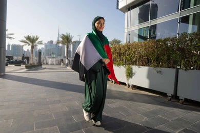 الاحتفال بذكرى استقلال دولة الإمارات العربية المتحدة، اليوم الوطني الإماراتي 2 ديسمبر، امراة عربية خليجية اماراتية ترتدي العباءة والحجاب تضع علم الإمارات على كتفيها في النهار، التجول في الشوارع بايماءات السعادة والفخر والاعتزاز، مفهوم حب الوطن والانتماء اليه