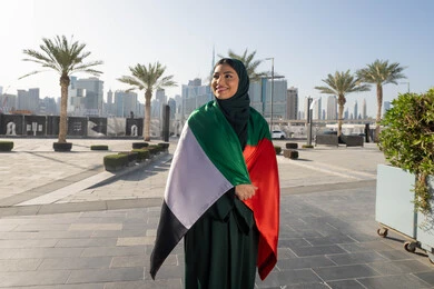 مفهوم حب الوطن والانتماء اليه، اليوم الوطني الإماراتي 2 ديسمبر، الاحتفال بذكرى استقلال دولة الإمارات العربية المتحدة، امراة عربية خليجية اماراتية ترتدي العباءة والحجاب تضع علم الإمارات على كتفيها في النهار، التجول في الشوارع بايماءات السعادة والفخر والاعتزاز