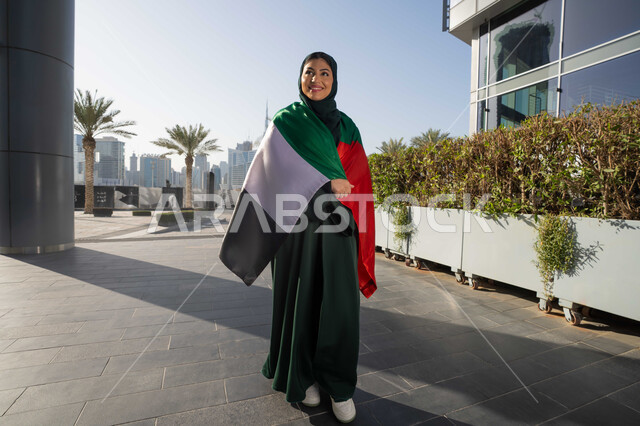 UAE National Day, December 2, celebrating the independence of the United Arab Emirates, an Emirati Gulf Arab woman wearing an abaya and hijab, placing the UAE flag on her shoulders during the day, walking in the streets with gestures of happiness, pride and honor, full body