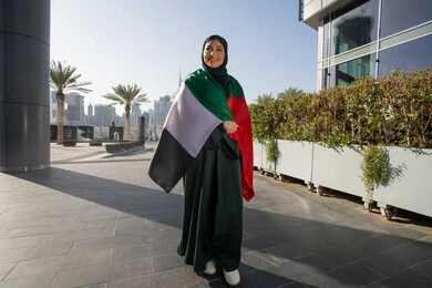 UAE National Day, December 2, celebrating the independence of the United Arab Emirates, an Emirati Gulf Arab woman wearing an abaya and hijab, placing the UAE flag on her shoulders during the day, walking in the streets with gestures of happiness, pride and honor, full body