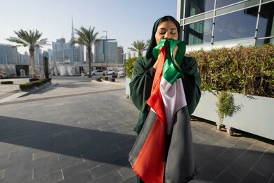 التجول في الشوارع بايماءات السعادة والفخر والاعتزاز، احياء اليوم الوطني الإماراتي 2 ديسمبر، الاحتفال بذكرى استقلال دولة الإمارات العربية المتحدة، امراة عربية خليجية اماراتية ترتدي العباءة والحجاب تقبل علم الإمارات في النهار، مفهوم حب الوطن والانتماء اليه