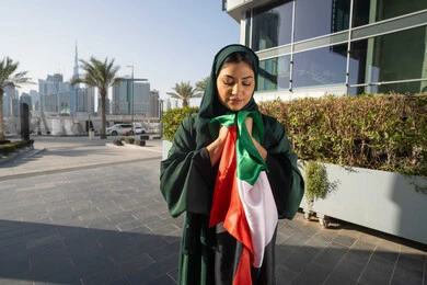 احياء اليوم الوطني الإماراتي 2 ديسمبر، الاحتفال بذكرى استقلال دولة الإمارات العربية المتحدة، امراة عربية خليجية اماراتية ترتدي العباءة والحجاب تقبل علم الإمارات في النهار، التجول في الشوارع بايماءات السعادة والفخر والاعتزاز، مفهوم حب الوطن والانتماء اليه