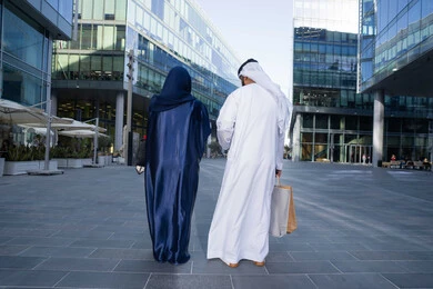 The pleasure of shopping in the malls of the United Arab Emirates, spending enjoyable times in nature, the concept of elegance and interest in external appearance, a picture from the back of an Arab Gulf Emirati couple wearing traditional dress carrying shopping bags, full body length