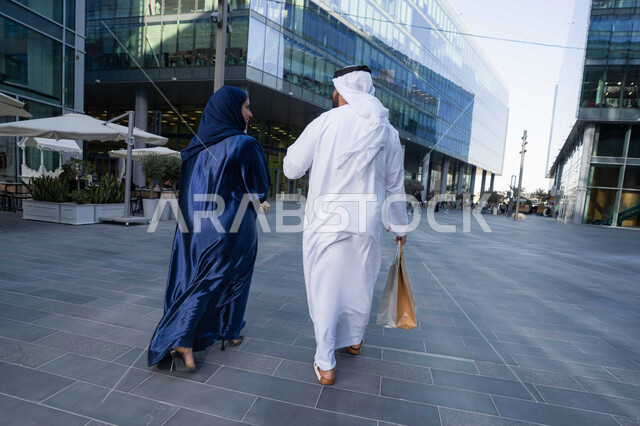 الاستمتاع والتجول واستنشاق الهواء النقي في وقت النهار، قضاء أوقات ممتعة في الطبيعة، مفهوم الأناقة والاهتمام بالمظهر الخارجي، صورة من الخلف لزوجان عربيان خليجيان اماراتيان يرتديان الزي التقليدي يحملان اكياس التسوق، الجسم بالطول الكامل