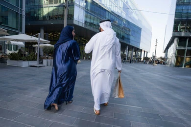 Enjoying, walking around and breathing fresh air during the day, spending enjoyable times in nature, the concept of elegance and interest in one's appearance, a photo from the back of an Emirati Gulf Arab couple wearing traditional dress carrying shopping bags, full body length