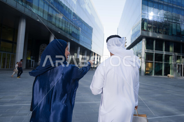 قضاء أوقات ممتعة في الطبيعة، مفهوم الأناقة والاهتمام بالمظهر الخارجي، الاستمتاع والتجول واستنشاق الهواء النقي في وقت النهار، صورة من الخلف لزوجان عربيان خليجيان اماراتيان يرتديان الزي التقليدي يحملان اكياس التسوق