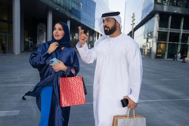الاستفادة من العروض الموسمية والتخفيضات، تعابير السرور والسعادة، التبضع وأخذ المستلزمات الضرورية، عروض الجمعه البيضاء، زوجان عربيان خليجيان اماراتيان يرتديان الزي التقليدي يحملان اكياس التسوق الملونه