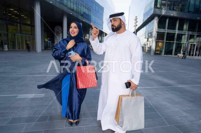 تعابير السرور والسعادة، التبضع وأخذ المستلزمات الضرورية، عروض الجمعه البيضاء، الاستفادة من العروض الموسمية والتخفيضات، زوجان عربيان خليجيان اماراتيان يرتديان الزي التقليدي يحملان اكياس التسوق الملونه، الجسم بالطول الكامل