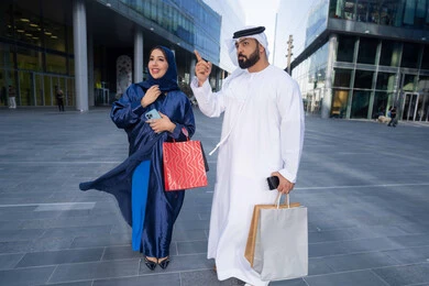 تعابير السرور والسعادة، التبضع وأخذ المستلزمات الضرورية، عروض الجمعه البيضاء، الاستفادة من العروض الموسمية والتخفيضات، زوجان عربيان خليجيان اماراتيان يرتديان الزي التقليدي يحملان اكياس التسوق الملونه، الجسم بالطول الكامل