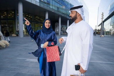 التبضع وأخذ المستلزمات الضرورية، تعابير السرور والسعادة، عروض الجمعه البيضاء، الاستفادة من العروض الموسمية والتخفيضات، زوجان عربيان خليجيان اماراتيان يرتديان الزي التقليدي يحملان اكياس التسوق الملونه