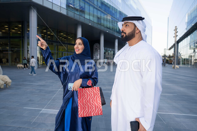 الاستمتاع بالشراء والتبضع، مشاركة الزوجين في اختيارات التسوق، قضاء أوقات ممتعة في الطبيعة ، الاستمتاع والتجول واستنشاق الهواء النقي في وقت النهار، زوجان عربيان خليجيان اماراتيان يرتديان الزي التقليدي يحملان اكياس التسوق بتعابير السرور