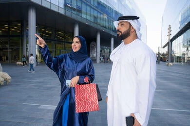 Enjoying shopping, sharing shopping choices with the couple, spending enjoyable times in nature, enjoying walking around and breathing fresh air during the day, an Arab Gulf Emirati couple wearing traditional dress carrying shopping bags with expressions of pleasure
