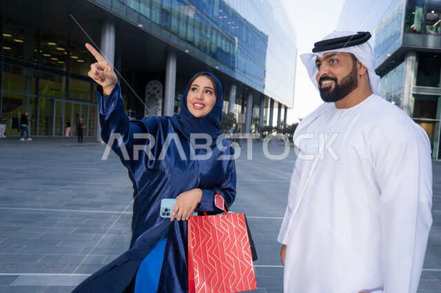 Taking advantage of seasonal offers and discounts, purchasing supplies and gifts, sharing shopping choices with the couple, spending enjoyable times in nature, enjoying walking around and breathing fresh air during the day, an Emirati Gulf Arab couple wearing traditional dress carrying shopping bags with happy expressions