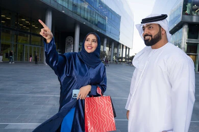 Taking advantage of seasonal offers and discounts, purchasing supplies and gifts, sharing shopping choices with the couple, spending enjoyable times in nature, enjoying walking around and breathing fresh air during the day, an Emirati Gulf Arab couple wearing traditional dress carrying shopping bags with happy expressions