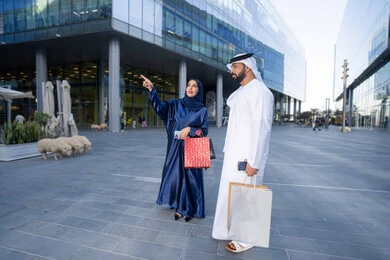 Buying necessities and gifts, sharing shopping choices with the couple, spending quality time in nature, enjoying walking around and breathing fresh air during the day, an Emirati Gulf Arab couple wearing traditional dress carrying shopping bags with happy expressions