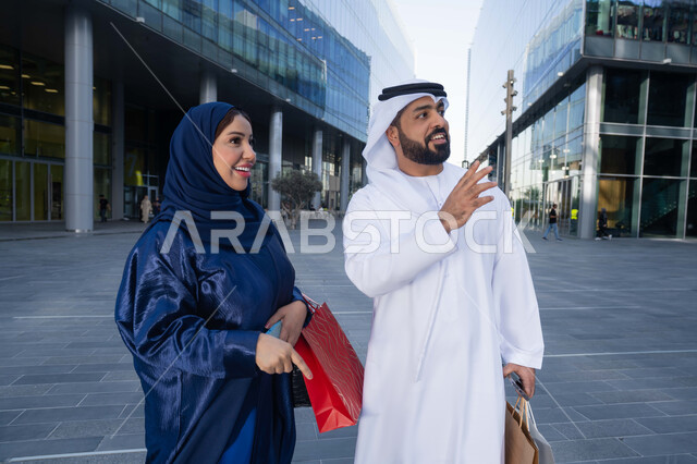 مشاركة الزوجين في اختيارات التسوق، قضاء أوقات ممتعة في الطبيعة ، الاستمتاع والتجول واستنشاق الهواء النقي في وقت النهار، زوجان عربيان خليجيان اماراتيان يرتديان الزي التقليدي يحملان اكياس التسوق بتعابير السرور