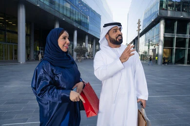 Couple sharing shopping choices, spending quality time in nature, enjoying walking around and breathing fresh air during the day, an Emirati Gulf Arab couple wearing traditional dress carrying shopping bags with happy expressions