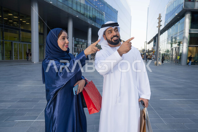 التسوق في المول، قضاء أوقات ممتعة في الطبيعة ، الاستمتاع والتجول واستنشاق الهواء النقي في وقت النهار، زوجان عربيان خليجيان اماراتيان يرتديان الزي التقليدي يحملان اكياس التسوق بتعابير السرور يقومان بالاشارة الى شي ما