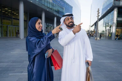Shopping in the mall, having fun in nature, enjoying walking around and breathing fresh air during the day, an Arab Gulf Emirati couple wearing traditional dress carrying shopping bags with happy expressions and pointing to something