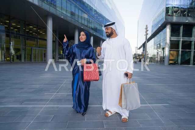 قضاء أوقات ممتعة في الطبيعة ، مفهوم الأناقة والاهتمام بالمظهر الخارجي ، الاستمتاع والتجول واستنشاق الهواء النقي في وقت النهار، زوجان عربيان خليجيان اماراتيان يرتديان الزي التقليدي يحملان اكياس التسوق بتعابير السرور، الجسم بالطول الكامل