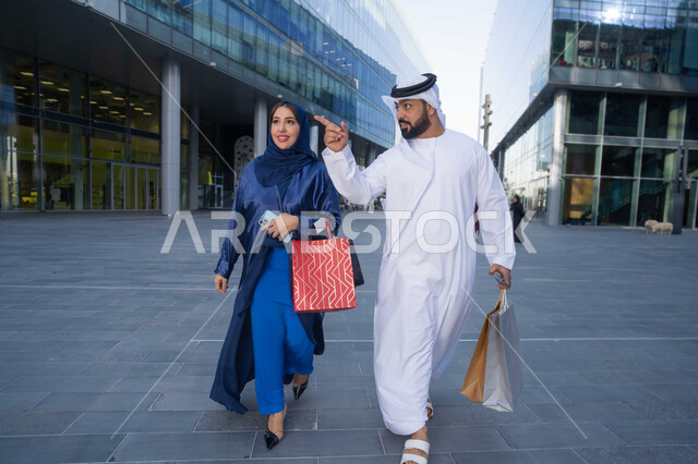 مفهوم الأناقة والاهتمام بالمظهر الخارجي ، قضاء أوقات ممتعة في الطبيعة ، الاستمتاع والتجول واستنشاق الهواء النقي في وقت النهار، زوجان عربيان خليجيان اماراتيان يرتديان الزي التقليدي يحملان اكياس التسوق بتعابير السرور، الجسم بالطول الكامل