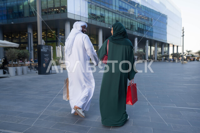 اقتناء الهدايا والحاجات الضرورية، صورة من الخلف لزوجان عربيان خليجيان اماراتيان يرتديان الزي التقليدي يحملان اكياس التسوق الملونه، التبضع وشراء المستلزمات من المولات التجارية في الامارت العربية، عروض الجمعه البيضاء