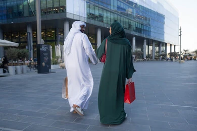 Buying gifts and essentials, a back view of an Emirati Gulf Arab couple wearing traditional dress carrying colorful shopping bags, shopping and buying supplies from malls in the UAE, White Friday offers