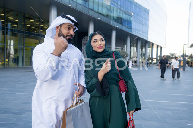 قضاء وقت ممتع في شراء المستلزمات، متعة التسوق في المول، زوجان عربيان خليجيان اماراتيان يرتديان الزي التقليدي يحملان اكياس التسوق الملونه، التبضع وشراء المستلزمات من المولات التجارية في الامارت العربية، عروض الجمعه البيضاء