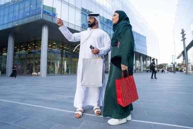 الاستمتاع بالشراء والتسوق ، الاستفادة من العروض الموسمية والتخفيضات، زوجان عربيان خليجيان اماراتيان يرتديان الزي التقليدي يحملان اكياس التسوق الملونه، التبضع وشراء المستلزمات من المولات التجارية في الامارت العربية، الجسم بالطول الكامل