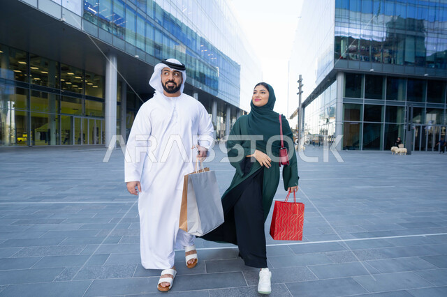 الأناقة والاهتمام بالمظهر الخارجي، الاستفادة من العروض الموسمية والتخفيضات، زوجان عربيان خليجيان اماراتيان يرتديان الزي التقليدي يحملان اكياس التسوق الملونه، التبضع وشراء المستلزمات من المولات التجارية في الامارت العربية، الجسم بالطول الكامل