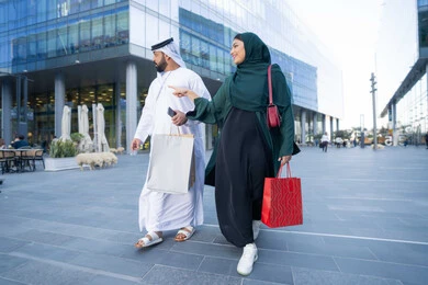 Business purchases and gifts, taking advantage of seasonal offers and discounts, an Arab Gulf Emirati couple wearing traditional dress carrying colorful shopping bags, shopping and buying supplies from shopping malls in the United Arab Emirates, full body