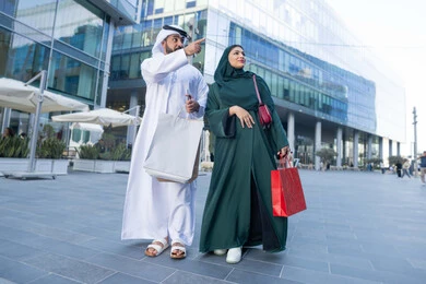Shopping and taking the necessary supplies, expressions of pleasure and happiness, Black Friday offers, taking advantage of seasonal offers and discounts, an Arab Gulf Emirati couple wearing traditional dress carrying colorful shopping bags, full body