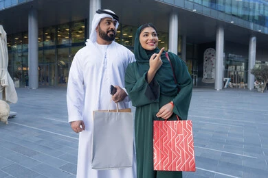 Buying groceries and gifts, expressions of joy and happiness, Black Friday offers, taking advantage of seasonal offers and discounts, an Arab Gulf Emirati couple wearing traditional dress carrying colorful shopping bags