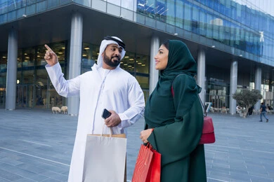 متعة التسوق واقتناء المستلزمات والهدايا من المحلات والمراكز التجاريه، تعابير السرور والسعادة، عروض الجمعه البيضاء، الاستفادة من العروض الموسمية والتخفيضات، زوجان عربيان خليجيان اماراتيان يرتديان الزي التقليدي يحملان اكياس التسوق الملونه