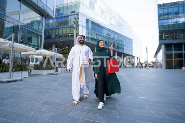 Shopping and purchasing supplies from shopping malls in the UAE, taking advantage of seasonal offers and discounts, an Emirati Gulf Arab couple wearing traditional dress carrying colorful shopping bags, White Friday offers