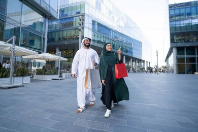 Shopping and purchasing supplies from shopping malls in the UAE, taking advantage of seasonal offers and discounts, an Emirati Gulf Arab couple wearing traditional dress carrying colorful shopping bags, White Friday offers