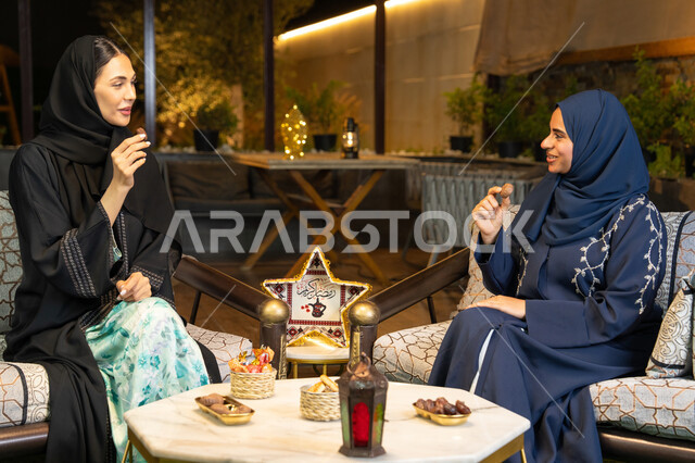 Eating fresh sugar dates, a local Emirati product, a warm welcome during the holy month of Ramadan, Ramadan decorations and lighting, two Saudi women sitting outdoors in a chalet in the Emirates, two Emirati Gulf Arab friends wearing the abaya sitting outdoors