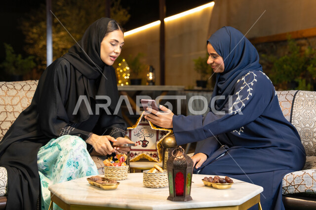 زينة واضاءة رمضانية، استخدام جهاز الهاتف المحمول، امراتان سعوديتان تجلسان بالجلسة الخارجية في احدى شاليهات الامارات، حسن الاستقبال في شهر رمضان المبارك، صديقتان عربيتان خليجيتان اماراتيتان ترتديان العباءة تجلسان في الهواء الطلق