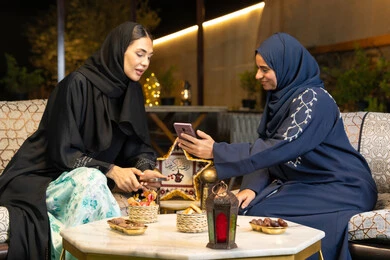 Ramadan decorations and lighting, using a mobile phone, two Saudi women sitting outdoors in a chalet in the Emirates, warm welcome in the holy month of Ramadan, two Emirati Gulf Arab friends wearing the abaya sitting outdoors