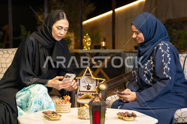 استخدام جهاز الهاتف المحمول، امراتان سعوديتان تجلسان بالجلسة الخارجية في احدى شاليهات الامارات، حسن الاستقبال في شهر رمضان المبارك، صديقتان عربيتان خليجيتان اماراتيتان ترتديان العباءة تجلسان في الهواء الطلق، زينة واضاءة رمضانية