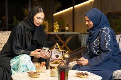 استخدام جهاز الهاتف المحمول، امراتان سعوديتان تجلسان بالجلسة الخارجية في احدى شاليهات الامارات، حسن الاستقبال في شهر رمضان المبارك، صديقتان عربيتان خليجيتان اماراتيتان ترتديان العباءة تجلسان في الهواء الطلق، زينة واضاءة رمضانية