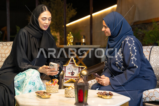 استخدام جهاز الهاتف المحمول، امراتان سعوديتان تجلسان بالجلسة الخارجية في احدى شاليهات الامارات، حسن الاستقبال في شهر رمضان المبارك، صديقتان عربيتان خليجيتان اماراتيتان ترتديان العباءة تجلسان في الهواء الطلق، زينة واضاءة رمضانية