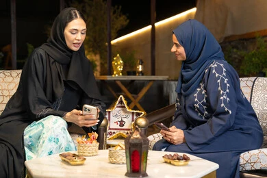 استخدام جهاز الهاتف المحمول، امراتان سعوديتان تجلسان بالجلسة الخارجية في احدى شاليهات الامارات، حسن الاستقبال في شهر رمضان المبارك، صديقتان عربيتان خليجيتان اماراتيتان ترتديان العباءة تجلسان في الهواء الطلق، زينة واضاءة رمضانية