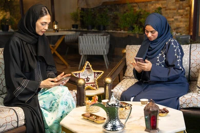 Warm welcome in the holy month of Ramadan, Ramadan decorations and lighting, using a mobile phone, two Saudi women sitting outdoors in a chalet in the Emirates, two Emirati Gulf Arab friends wearing the abaya sitting outdoors