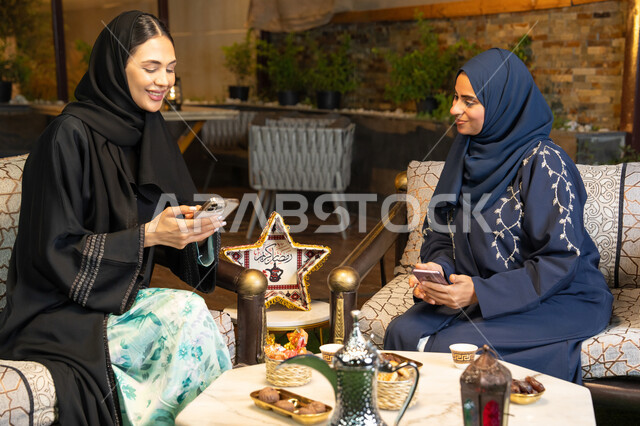 اضواء و زينة رمضانية، استخدام جهاز الهاتف المحمول، امراتان سعوديتان تجلسان بالجلسة الخارجية في احدى شاليهات الامارات، حسن الاستقبال في شهر رمضان المبارك، صديقتان عربيتان خليجيتان اماراتيتان ترتديان العباءة تجلسان في الهواء الطلق