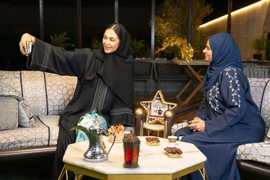 التقاط صور سيلفي باستخدام الهاتف المحمول، امراتان سعوديتان تقومان بتوثيق اللحظات السعيدة، حسن الاستقبال وكرم الضيافة في شهر رمضان المبارك، صديقتان عربيتان خليجينان اماراتيتان ترتديان العباءة تجلسان في الهواء الطلق، زينة واضاءة رمضانية، الجلسة الليلة الخارجية